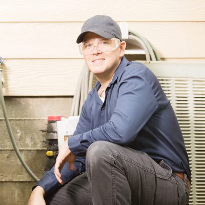 Mid-adult repairmen work on a home's air conditioner unit outdoors. Man center is working on the unit using tools in his toolbox.  Other man to right. They both wear uniforms.